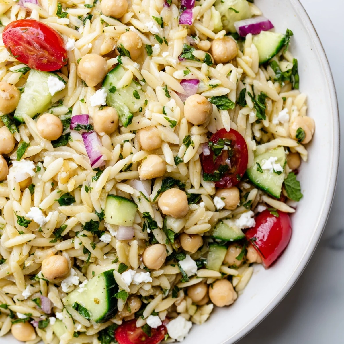 A bright bowl of Lemony Chickpea Orzo Salad with chickpeas, cucumbers, tomatoes, and herbs glistening in lemon vinaigrette.