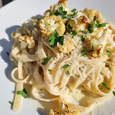 A skillet of hot, silky Roasted Cauliflower Alfredo sauce coats freshly cooked fettuccine, garnished with parsley and Parmesan.